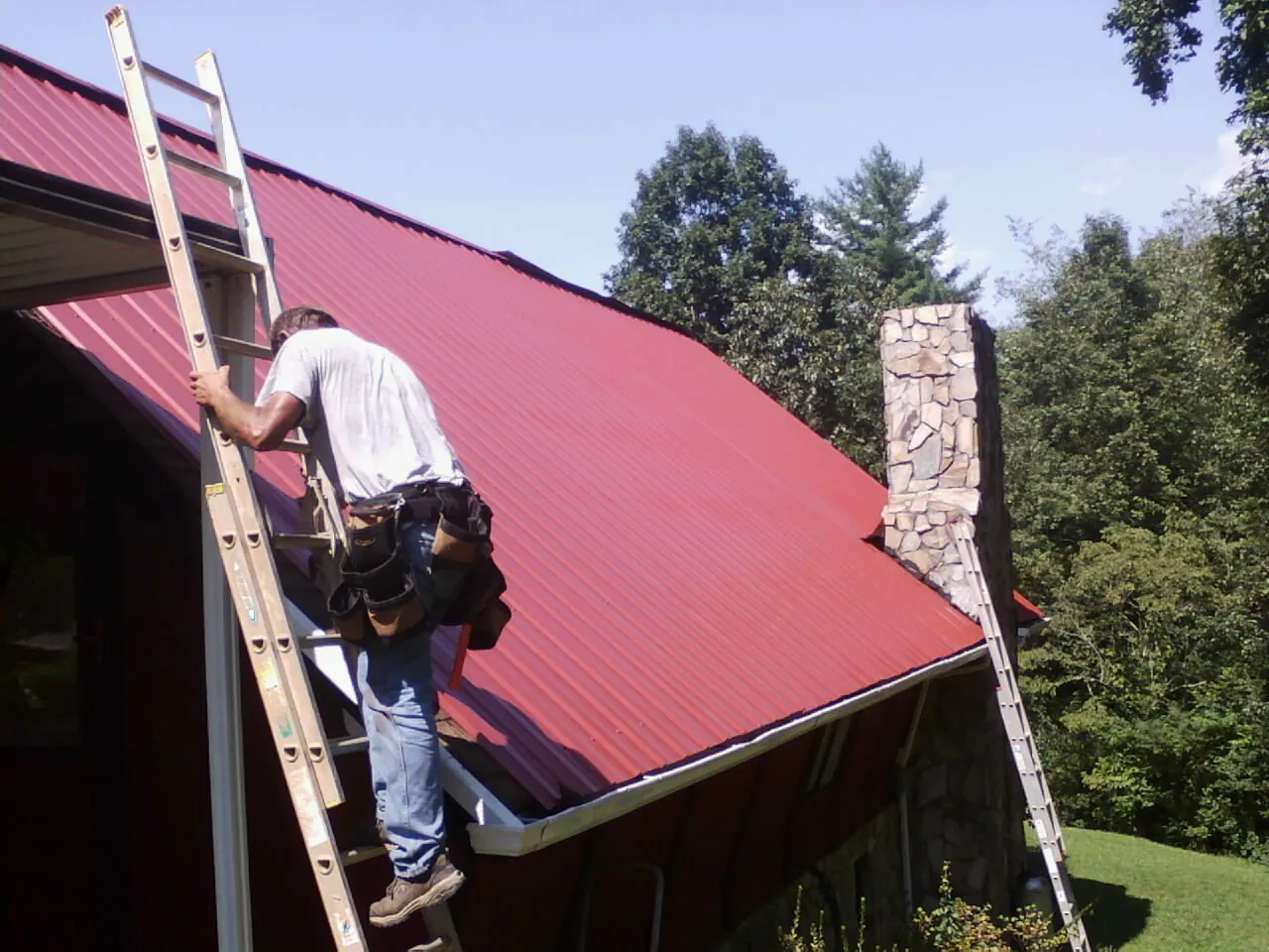 Roofer on a ladder beside a red metal roof and stone chimney — Soffit & Fascia Repair by Roofing Brothers in Batson